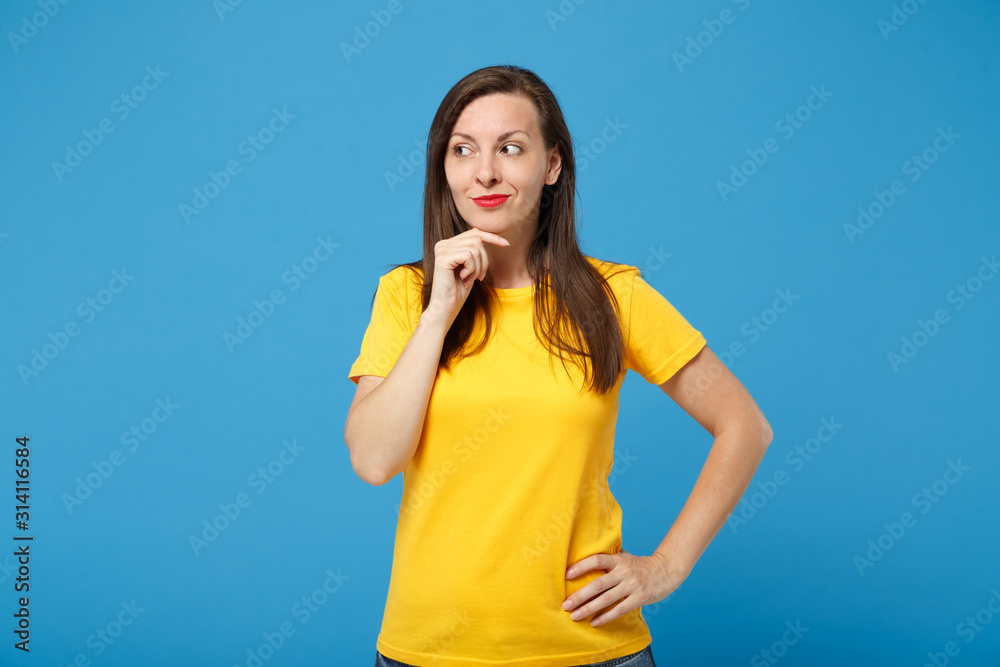 Smiling young brunette woman girl in yellow t-shirt posing isolated on bright blue background, studio portrait. People lifestyle concept. Mock up copy space. Looking aside, put hand prop up on chin. Smiling young brunette woman girl in yellow t-shirt posing isolated on bright blue background, studio portrait. People lifestyle concept. Mock up copy space. Looking aside, put hand prop up on chin.