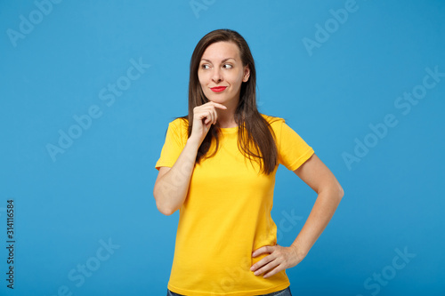 Smiling young brunette woman girl in yellow t-shirt posing isolated on bright blue background, studio portrait. People lifestyle concept. Mock up copy space. Looking aside, put hand prop up on chin.
