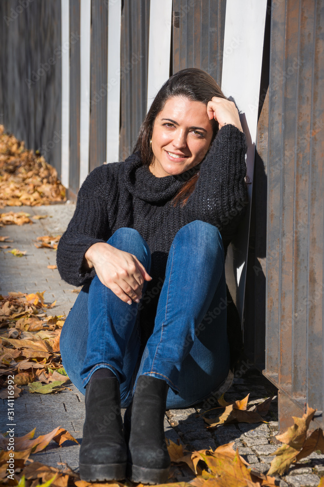 Smiling young brunette woman, sitting on the floor with fallen leaves, posing with casual winter clothes, a sunny day in Madrid, Spain, Europe