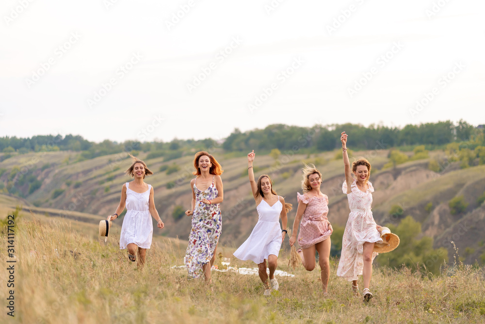 Obraz premium The company of cheerful female friends have a great time together on a picnic in a picturesque place overlooking the green hills. Girls in white dresses dancing in the field