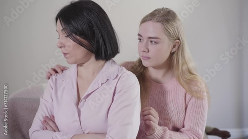 Young blond Caucasian girl touching mother's shoulder and smiling. Adult brunette woman turning to teen daughter and hugging her. Family making peace indoors. photo