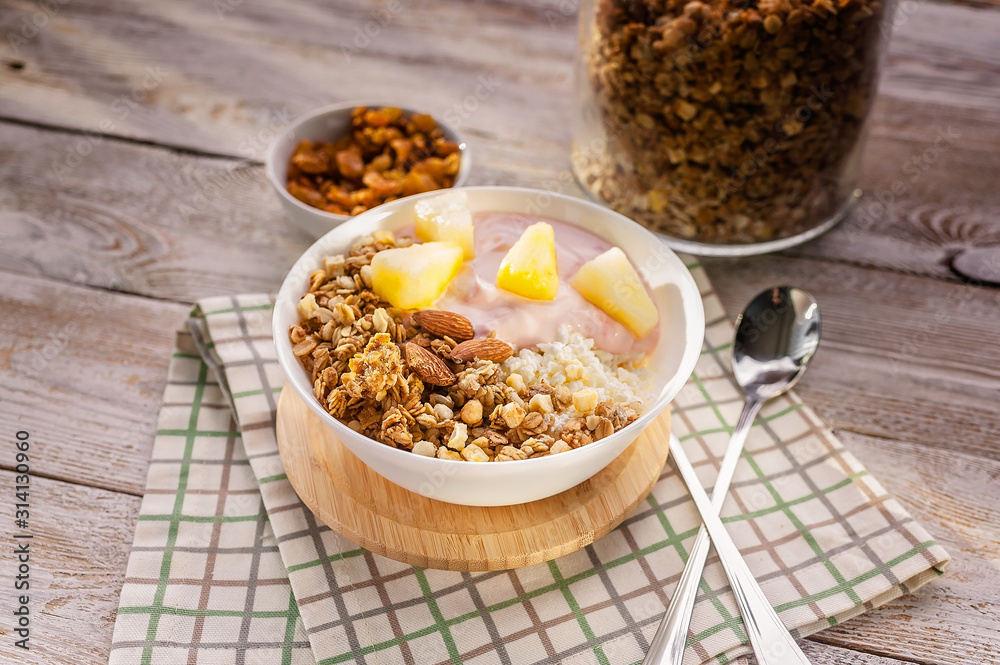Fototapeta premium Close up baked granola with nuts, cottage cheese, strawberry yogurt and pineapple slices in a plate on a checkered napkin. The light of the morning sun. Healthy breakfast