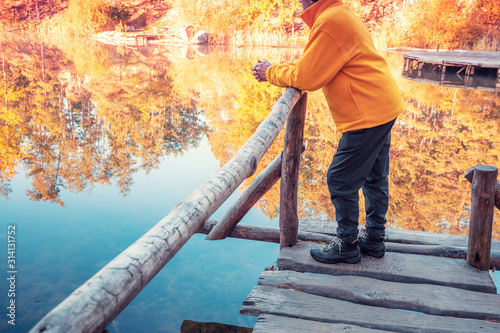 A man stands on a wooden deck leaning on a railing and looks at a forest lake. Sunrise over the lake in autumn