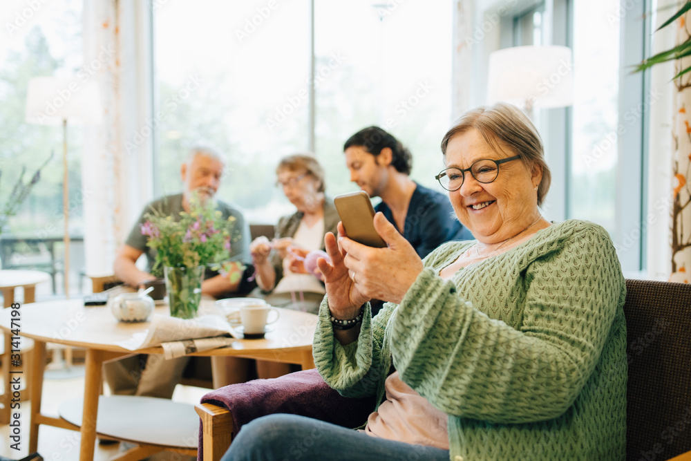 Smiling retired senior woman using smart phone while sitting on chair with friends and caregiver in background Smiling retired senior woman using smart phone while sitting on chair with friends and caregiver in background
