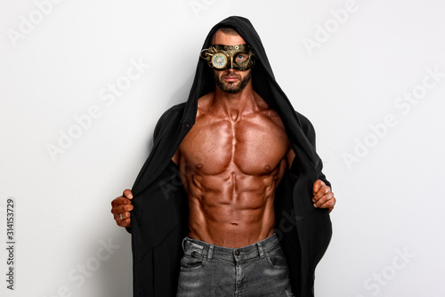 Handsome masked macho man posing  in unbuttoned coat with hood exposing his muscular build, in studio on white background photo