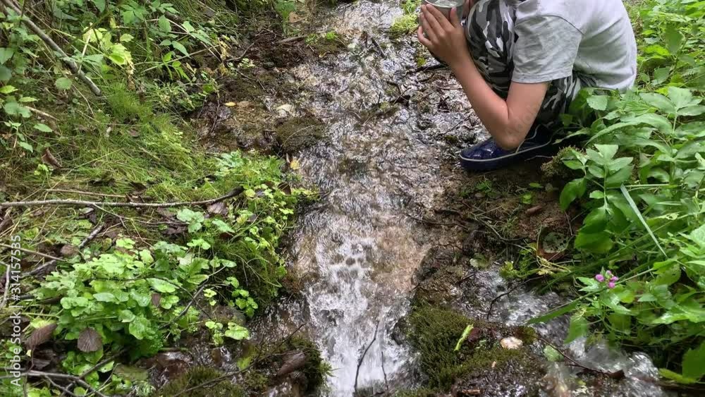 A boy hiker, trekker getting clear fresh water from a mountain spring in metal tourist cup and drinking A boy hiker, trekker getting clear fresh water from a mountain spring in metal tourist cup and drinking