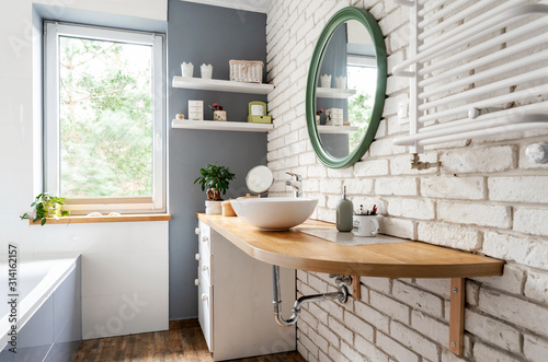 Tableau sur toile Bright interior of bathroom with window, wooden furniture and counter, round mirror and white brick wall