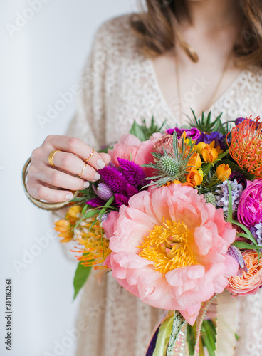 Young bride girl dressed in a dress holds a bouquet of natural flowers with multi-colored silk ribbons. Pink peonies, orange tulips, red roses. Boho chic style.