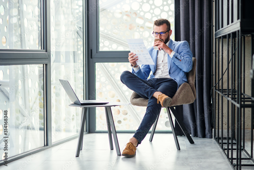 Young successful businessman analyzes financial document in his hand and drinks fragrant coffee in specially designated room in modern coworking centre. Young successful businessman analyzes financial document in his hand and drinks fragrant coffee in specially designated room in modern coworking centre.