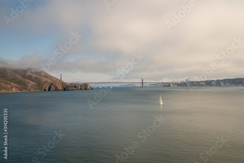 Yacht sailing in front of bridge with mountain cliffs on one side and cityscape skyline