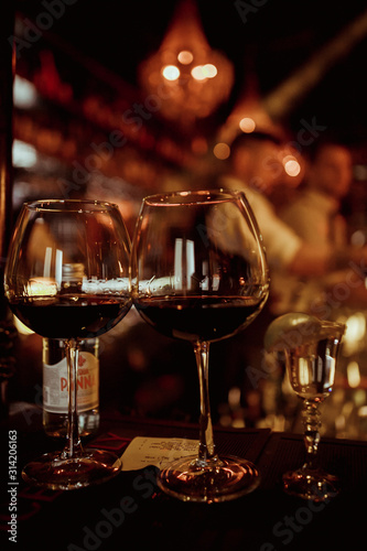 Low angle close up perspective of crystal clear wine glass with traditional round goblet shape filled with dark red wine and slim stem on wood counter top bar with blurry restaurant background scene