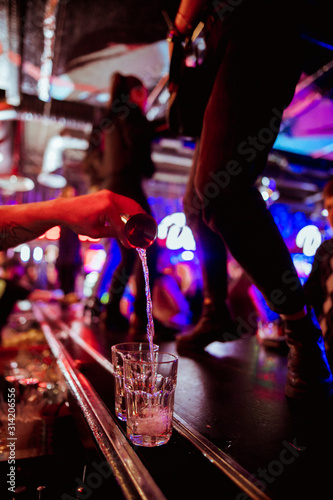 Bartender pouring a brown alcoholic cocktail from the measuring cup to the glass on the bar counter in the dark blurred background