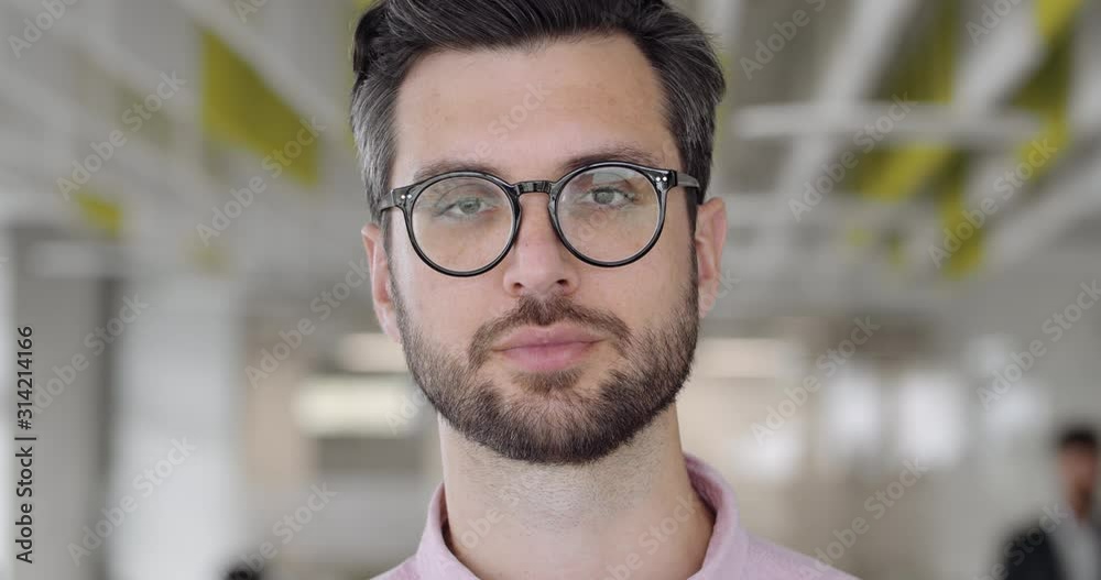 Close up of bearded caucasian man in 30s looking to camera and blinking. Portrait of handsome guy with serious face expression. Blurred modern office background. Concept of people.