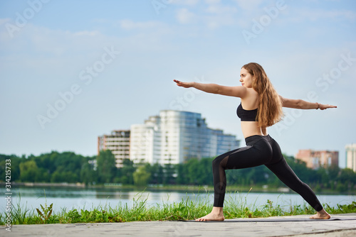 Yoga female with long hair standing in a warrior pose, Virabhadrasana near a river in the background of urban buildings and a park under a blue sky with copy space. Sport and healthy lifestyle concept
