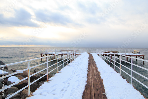 Winter beach. Blue clear sea water  snow and a wooden pier. Skyline and mountains.