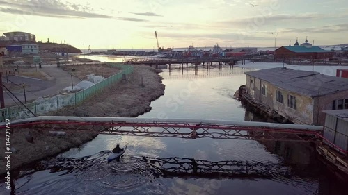 active canoeist silhouette sails under metal gas pipe bridge along river reflecting evening sunlight aerial backside view