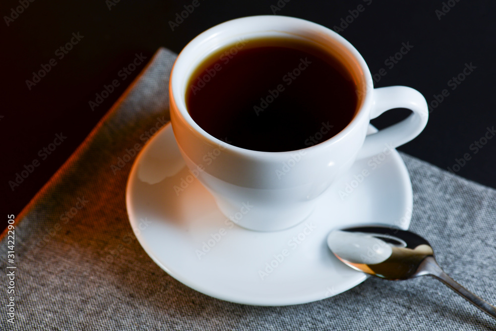 White cup of black tea served on a plate with a spoon on dark table background. Hot drink. White cup of black tea served on a plate with a spoon on dark table background. Hot drink.
