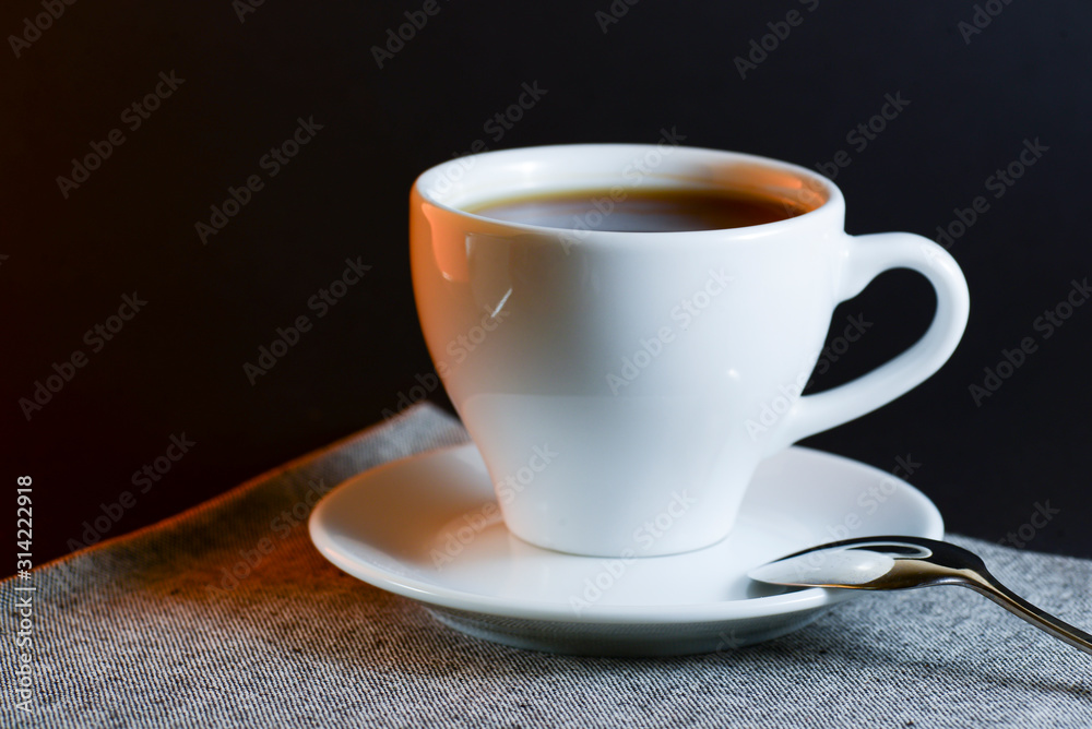 White cup of black tea served on a plate with a spoon on dark table background. Hot drink.