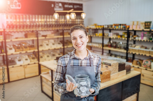 Mixed Race Woman Owner Selling Superfoods in Zero Waste Shop. Lots of Healthy Food in Glass Bottles on Stand in Grocery Store. No plastic Conscious Minimalism Vegan Lifestyle Concept. Selective focus