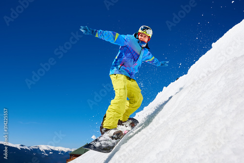 Snowboarder man riding snowboard fast down steep snowy mountain slope, jumping in air on copy space background of blue sky and white snow on sunny winter day. Extreme sport and recreation concept.