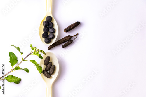 Top view of Black and brown ayurvedic medicine tablets in two spoons with dry long pepper and ocimum sanctum shoots on white background. Herbal medicine concept photo