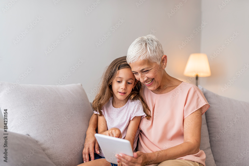 Smiling granddaughter and grandmother with tablet pc computer sitting on couch at home. Picture of cheerful young lady sitting at home with her grandmother using tablet computer. Smiling granddaughter and grandmother with tablet pc computer sitting on couch at home. Picture of cheerful young lady sitting at home with her grandmother using tablet computer.