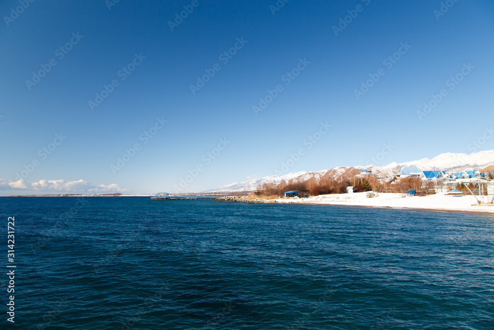 Winter beach. Blue clear sea water, snow and beach umbrellas. Skyline and mountains. Kyrgyzstan, Issyk-Kul Lake