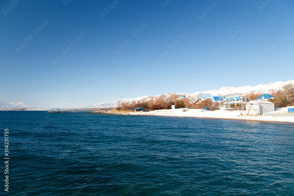 Winter beach. Blue clear sea water, snow and beach umbrellas. Skyline and mountains. Kyrgyzstan, Issyk-Kul Lake