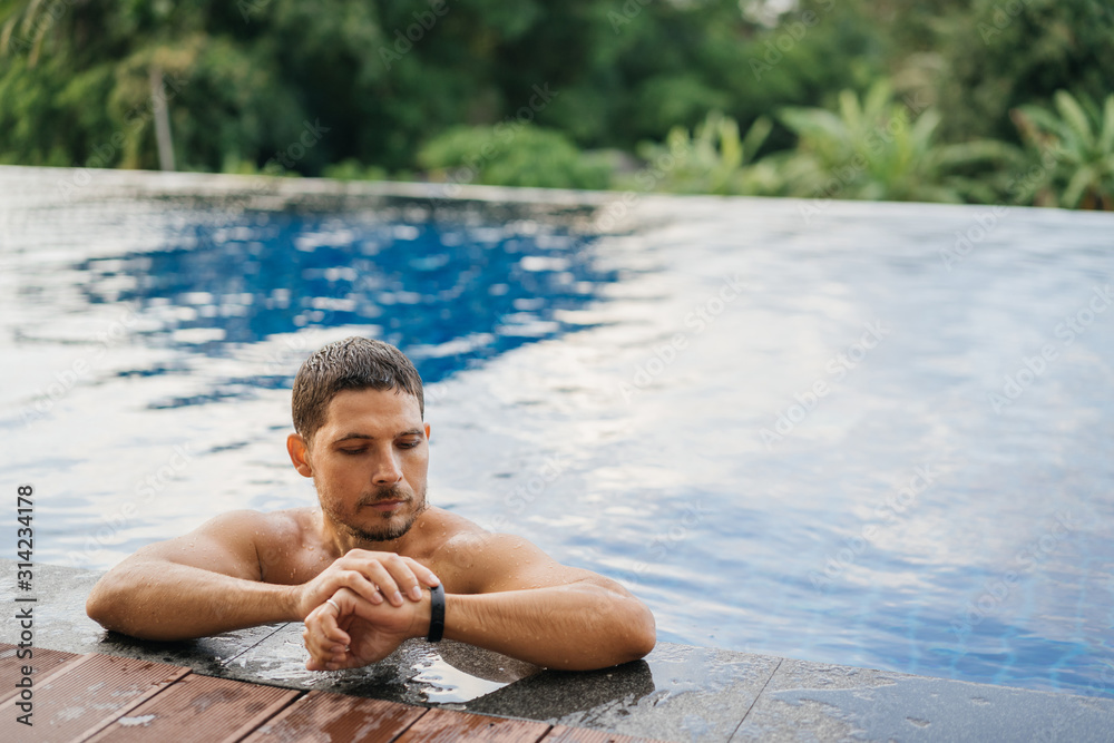 Fototapeta premium An attractive sporty man in an outdoor pool looks at the smartwatch tracker after an active swim.