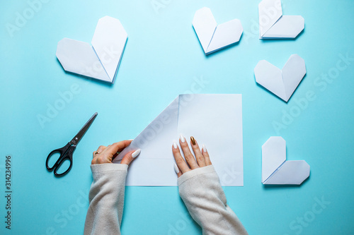 Many white origami hearts on blue background with sheet of paper and scissors in the center. Saint Valentine's day card with woman making origami heart on blue background.