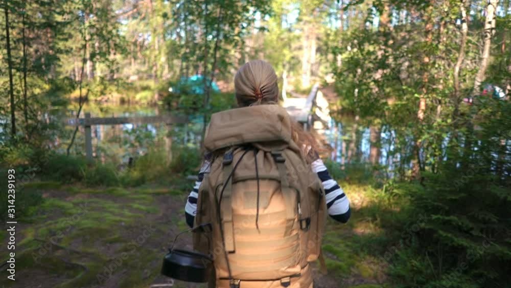 A young woman with a large backpack hiking a tourist forest trail on a bridge over a river in Finland