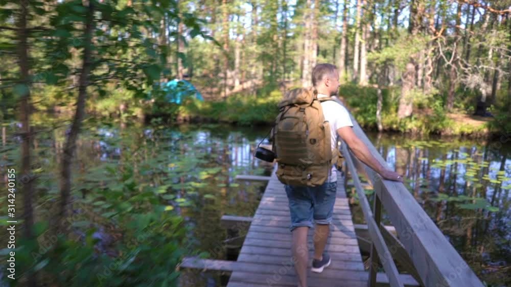 A young man with a large backpack hiking a tourist forest trail on a bridge over a river in Finland A young man with a large backpack hiking a tourist forest trail on a bridge over a river in Finland