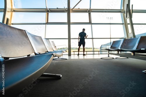 Young man holding smartphone in hands while standing in airport terminal. Focus on bench.