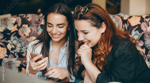 Two lovely young female looking at a smartphone screen laughing while sitting in a coffee shop.