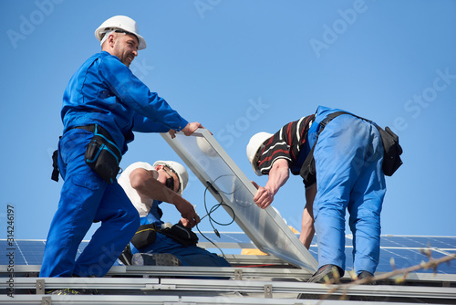 Male workers installing stand-alone solar photovoltaic panel system. Electricians mounting blue solar module on roof of modern house. Alternative energy ecological concept.