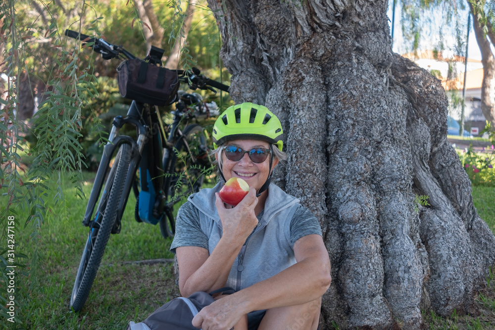 Attractive senior woman cyclist with yellow helmet have a break in the park near an old and big log. Moment of relaxation eating a red apple fruit. Green lawn and plants around her.