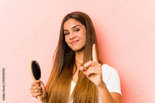 Young caucasian woman holding an hairbrush showing number one with finger.