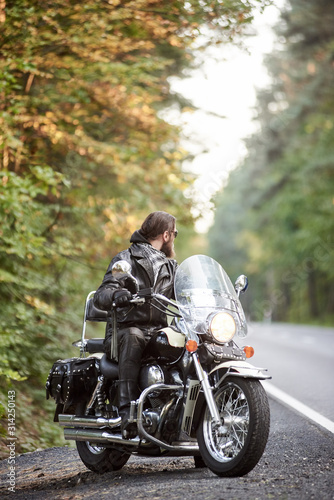 Bearded biker in black leather clothing sitting on cruiser motorcycle on country roadside on background of empty straight asphalt road and green trees bokeh foliage.