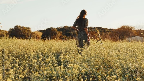 Girl in green dress walks walks through the fields fool of blooming yellow flowers. Long hair woman walks around the beautiful countryside. Golden light in idyllic landscape. Peace and tranquility.