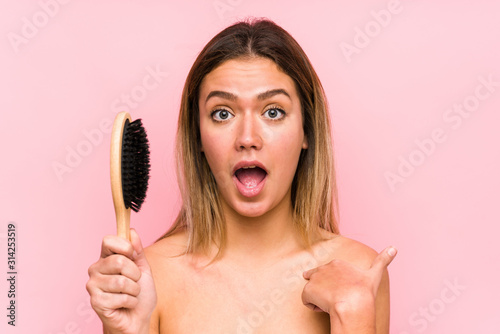 Young caucasian woman holding a hairbrush isolated surprised pointing at himself, smiling broadly.