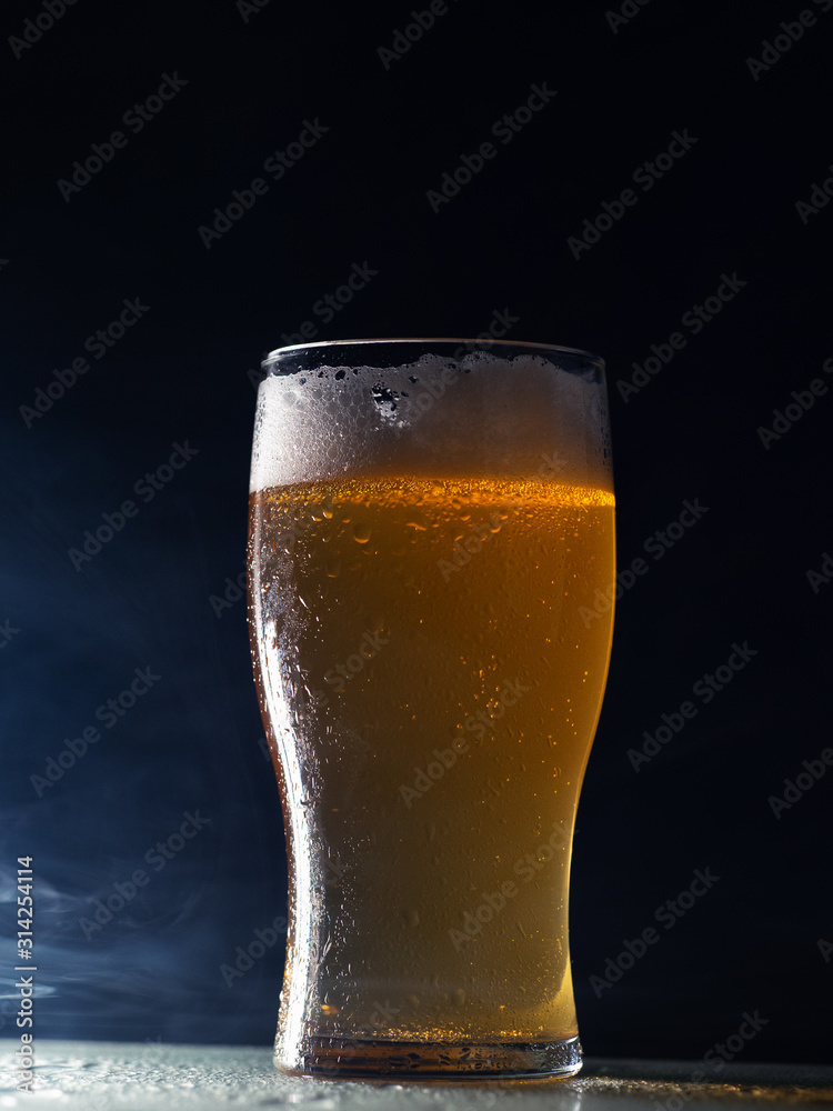 A glass of cold light beer lager on a dark background with smoke
