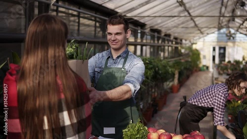 Portrait of european salesman wearing green apron is giving organic food in brown paper bag to female customer in greenhouse. Cheerful, smillling man selling organic food from the table while his wife photo