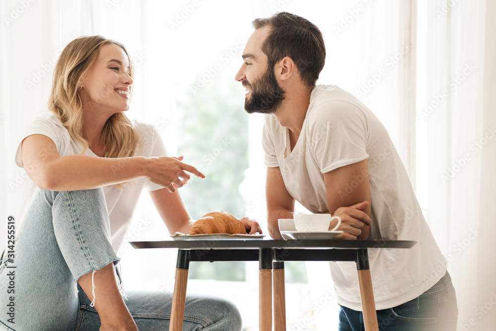 Happy beautiful young couple having breakfast at the kitchen table - obrazy, fototapety, plakaty 