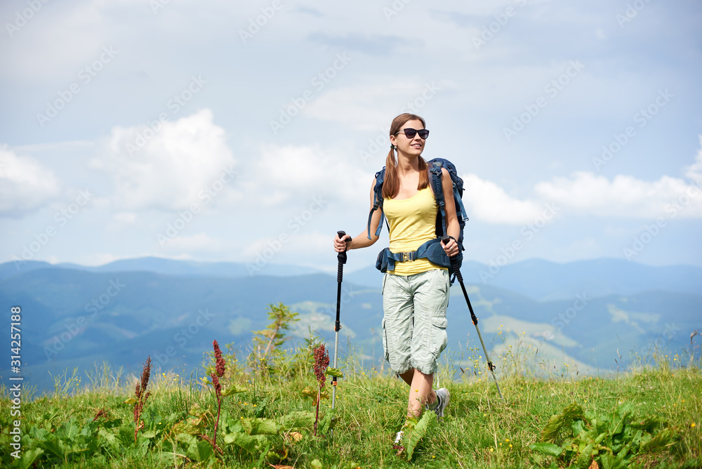 Athlete happy woman backpacker hiking mountain trail, walking on grassy hill, wearing backpack and sunglasses, using trekking sticks, enjoying summer cloudy day. Outdoor activity, tourism concept
