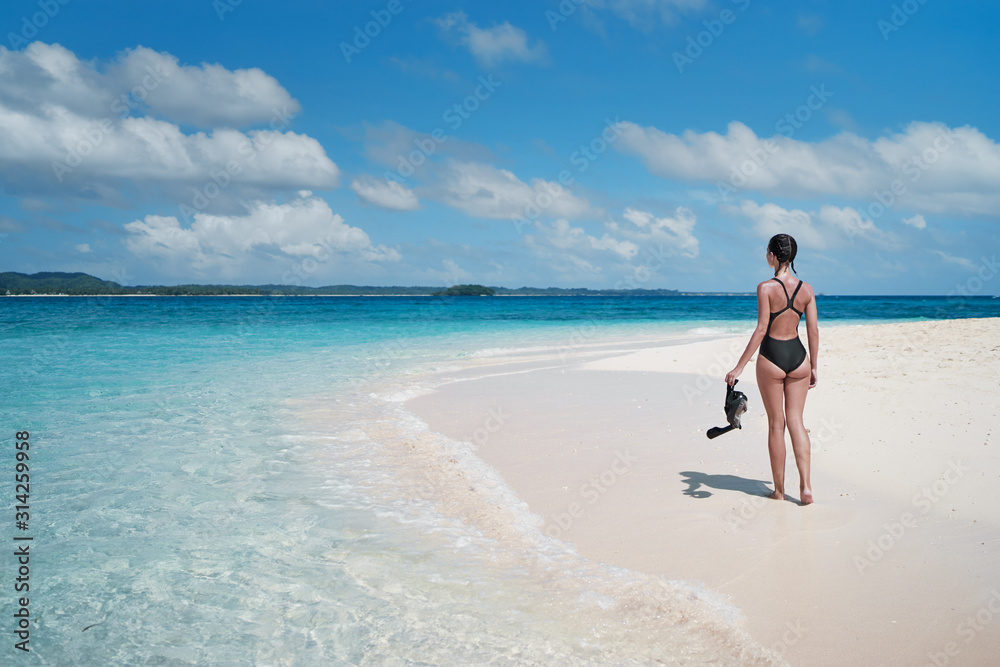 Tropical vacation. Young woman walking on tropical blue sea beach with snorkeling gear at sunny day.