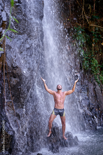 Happy young man relaxing under tropical waterfall with arms up raised in freedom. Health and relaxation.