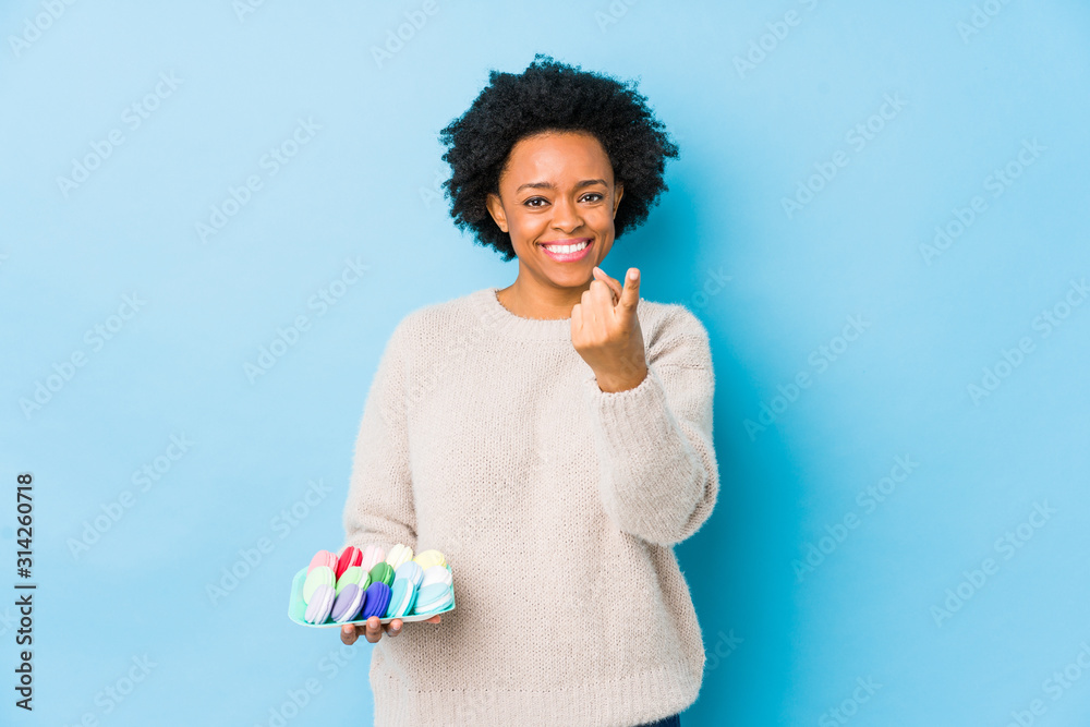 Middle age african american woman eating macaroons isolated pointing with finger at you as if inviting come closer. Middle age african american woman eating macaroons isolated pointing with finger at you as if inviting come closer.