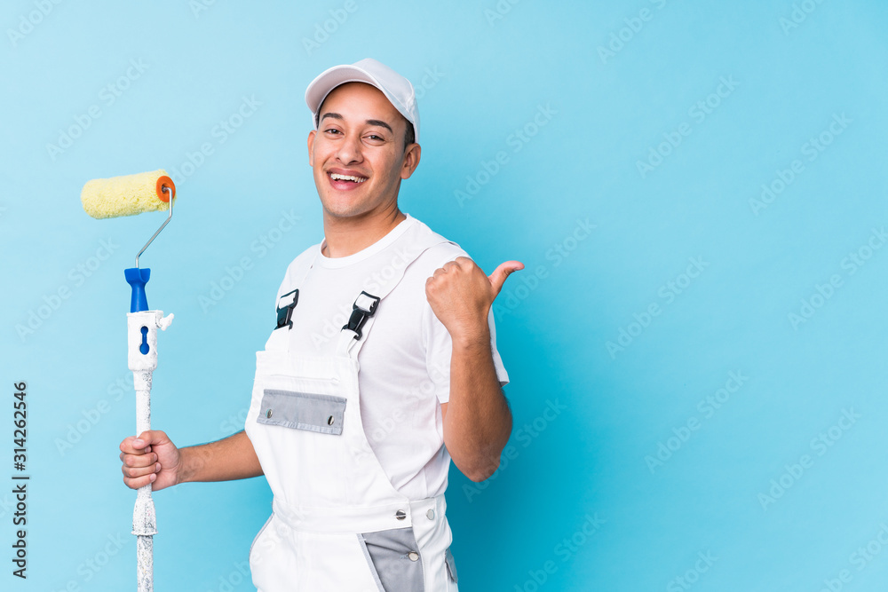 Young professional painter latin man isolated points with thumb finger away, laughing and carefree. - obrazy, fototapety, plakaty 