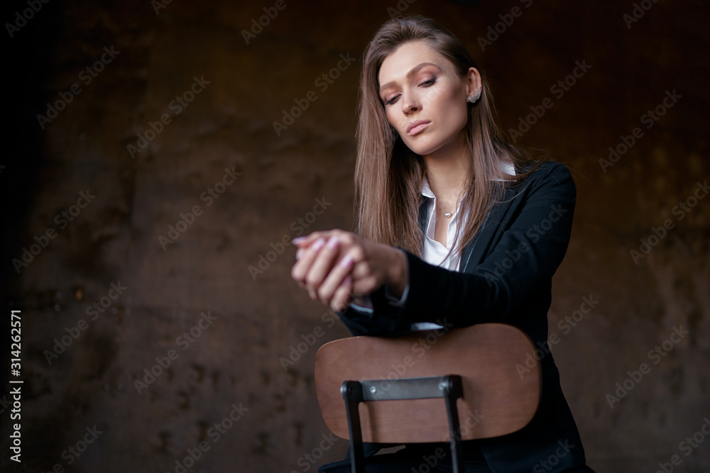 Studio portrait of young beautiful sensual woman in blak suit sitting on wooden chair against dark background.