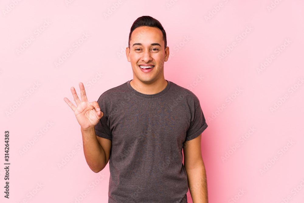 Yooung latin man posing in a pink backgroundwinks an eye and holds an okay gesture with hand.